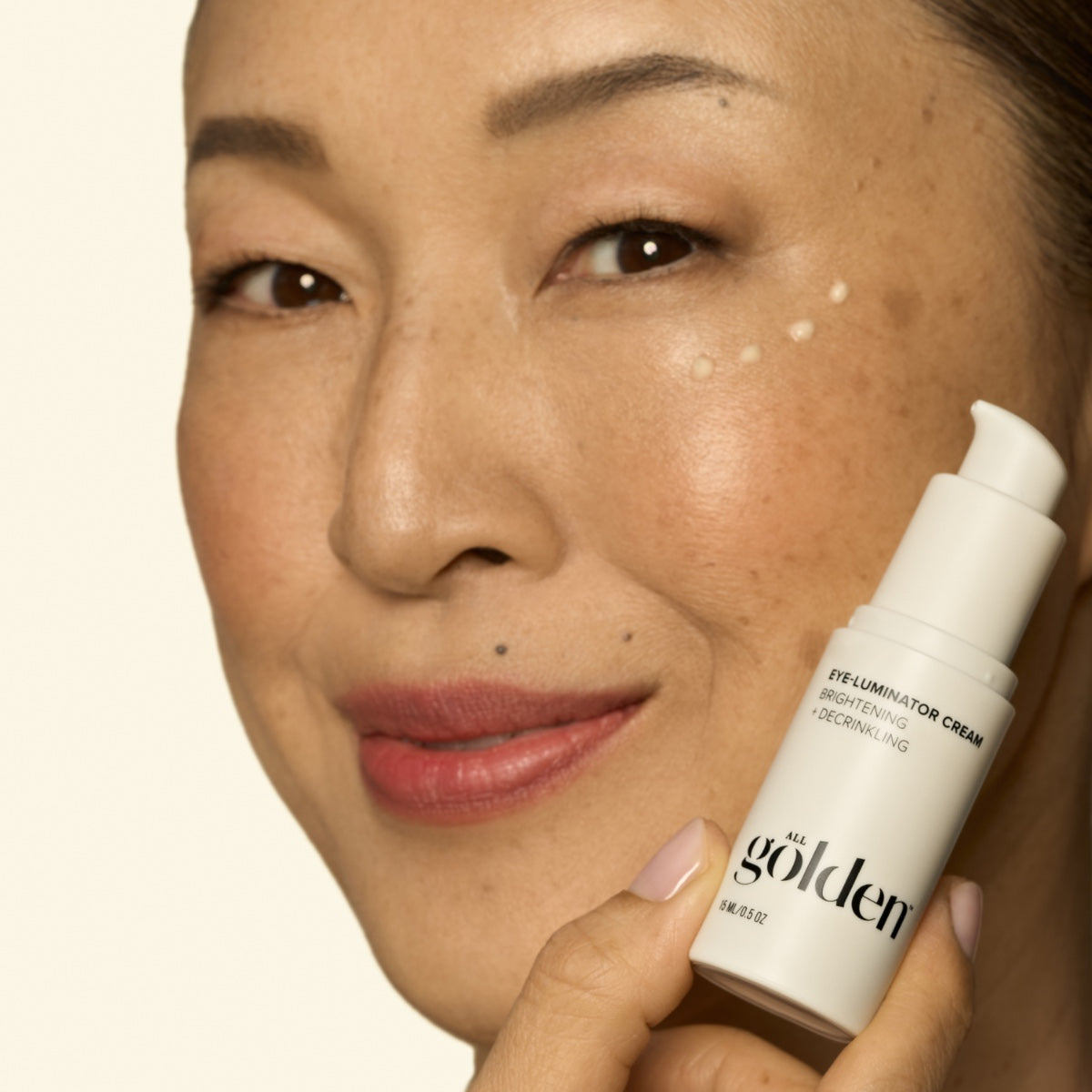 All Golden brightening under eye cream applied in dots on woman’s cheek near eyes with pump bottle