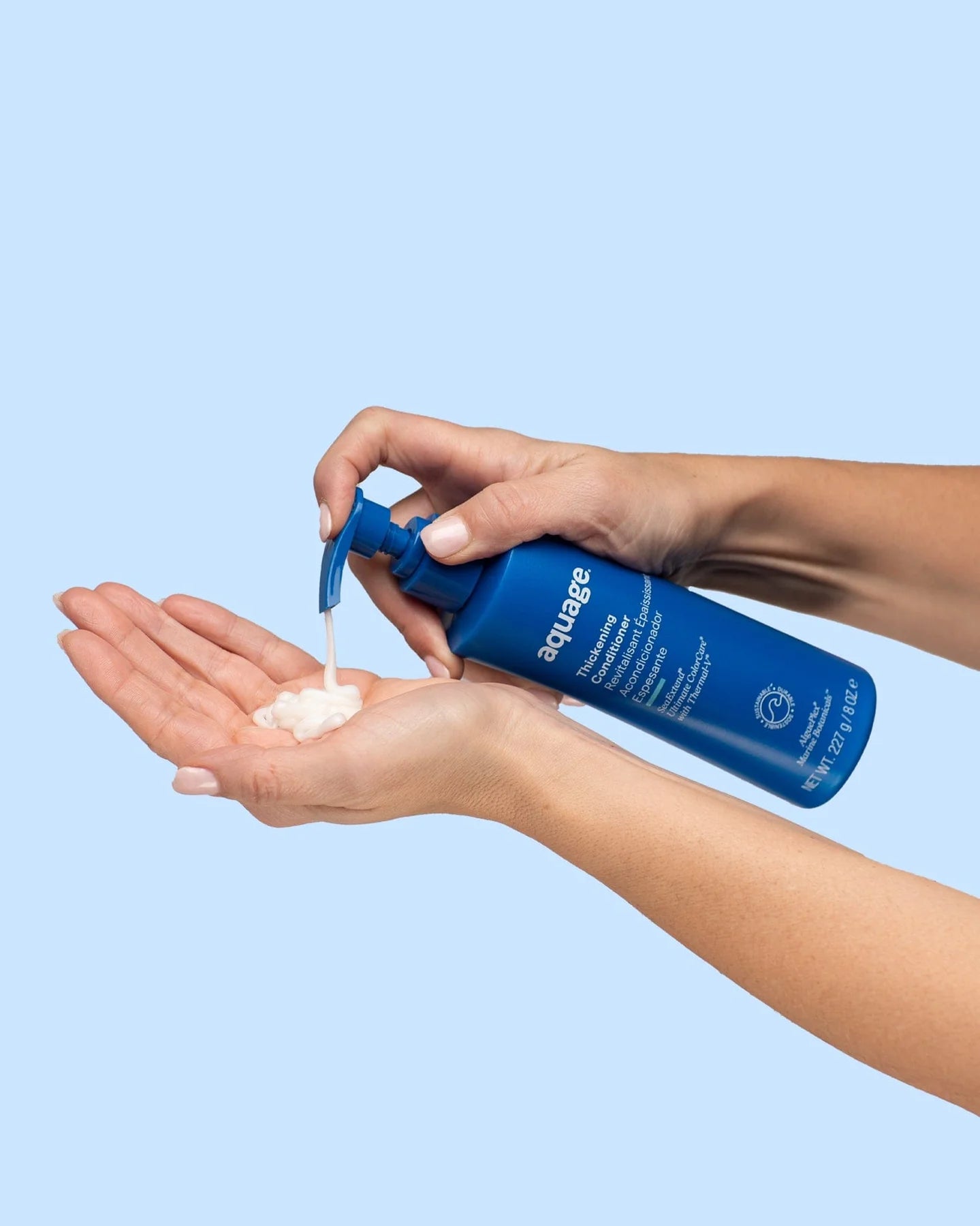 Hands dispensing white cream from blue bottle of Aquage thickening conditioner against light blue background