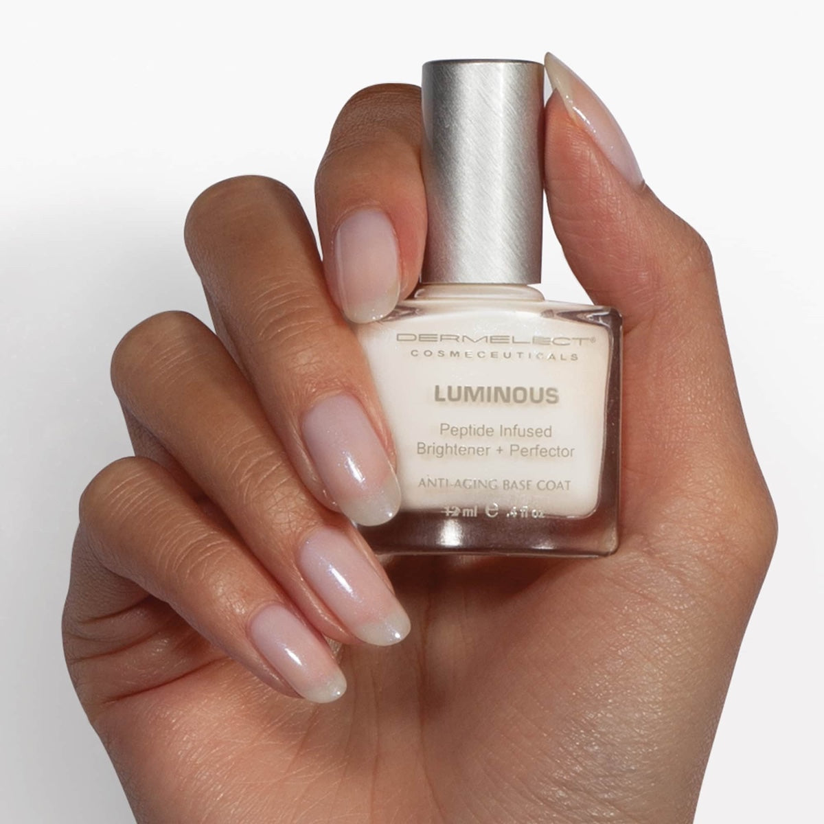 Hand with glossy natural nails holding Dermelect Luminous bottle anti aging base coat Dermelect Luminous Brightener review