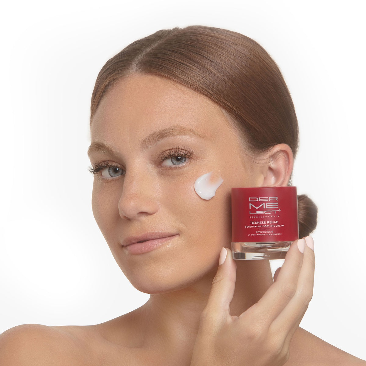 Smiling woman holds Dermelect Redness Rehab cream jar with white moisturizer dab on cheek