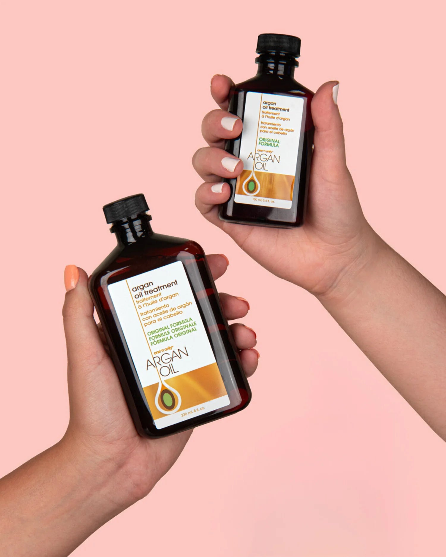 Two hands holding One N Only Argan Oil treatment bottles with black caps on pink background