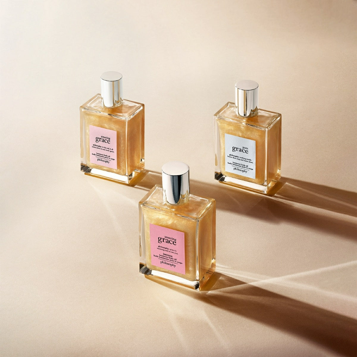 Three bottles of Philosophy Amazing Grace body oil with silver caps on a beige surface casting soft shadows