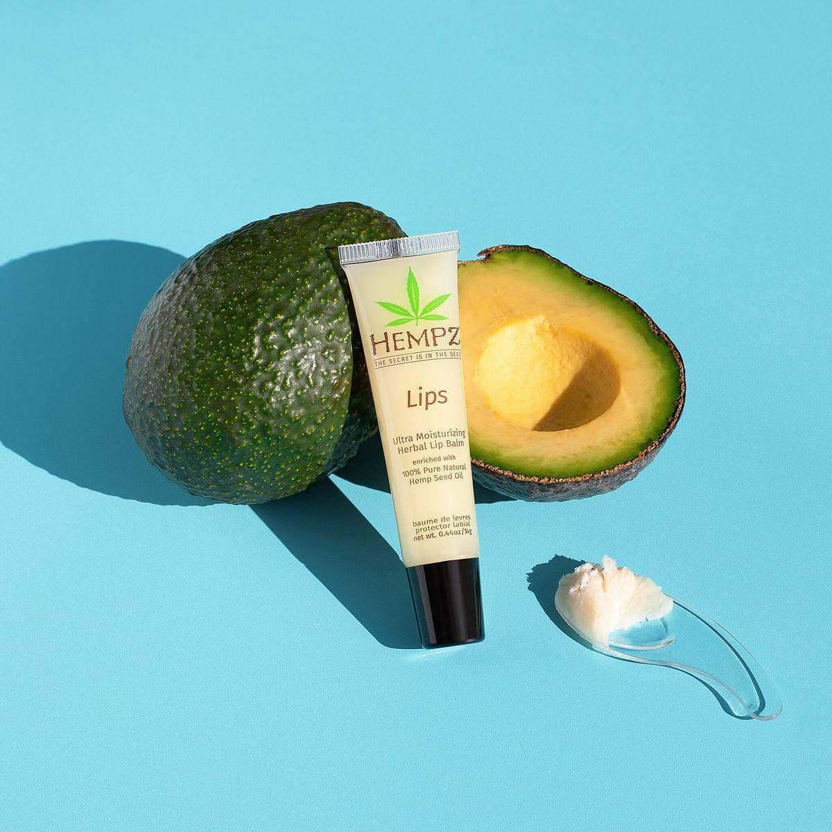 Hempz hemp lip balm tube with half avocado and spatula on blue background for moisturizing lips