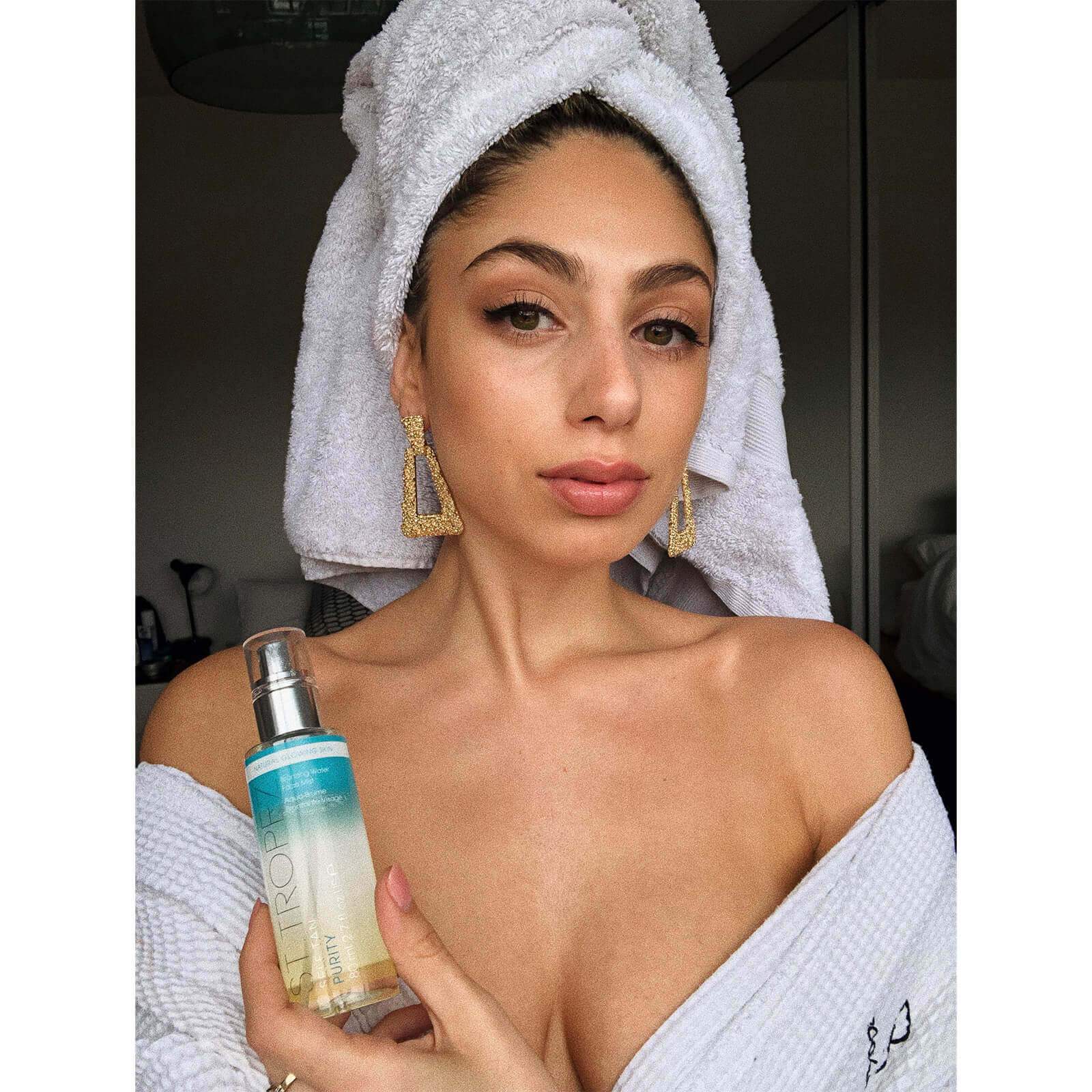 Woman in white robe and towel holding St. Tropez self tanner kit bottle with natural glowing skin