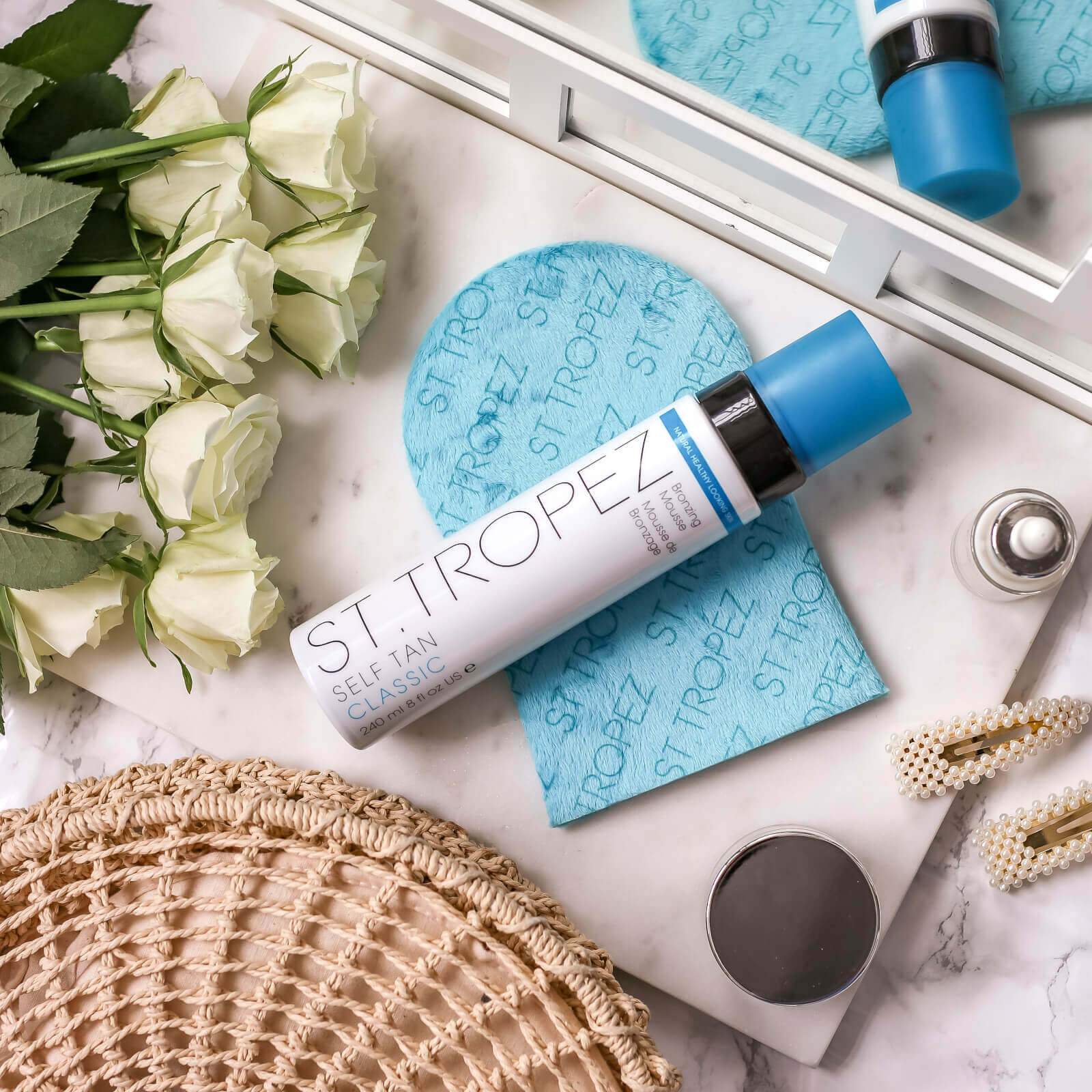 St Tropez self tanning mousse bottle with applicator mitt on marble surface near white roses and accessories