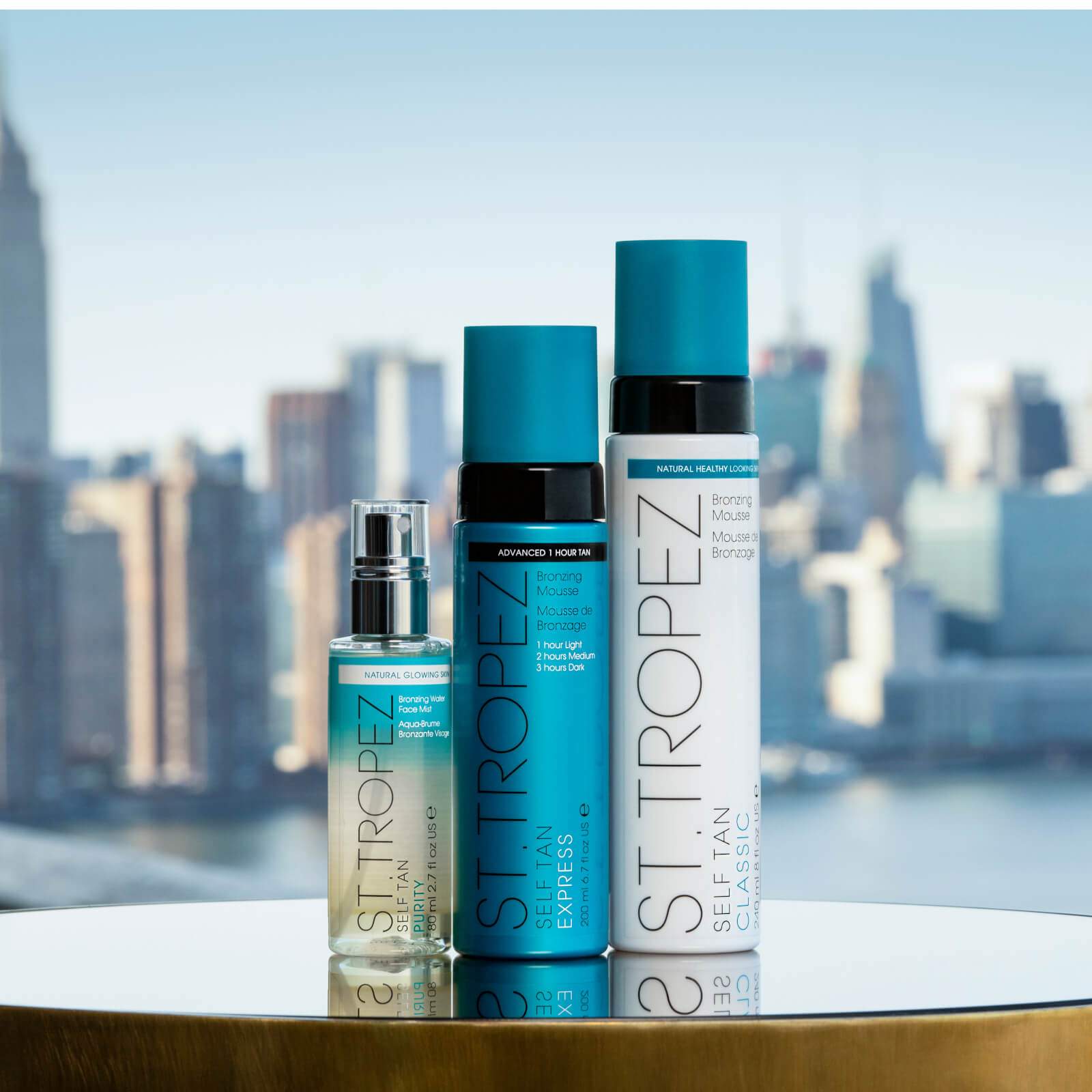 Three bottles of St Tropez self tanning mousse in different sizes and packaging with city skyline background