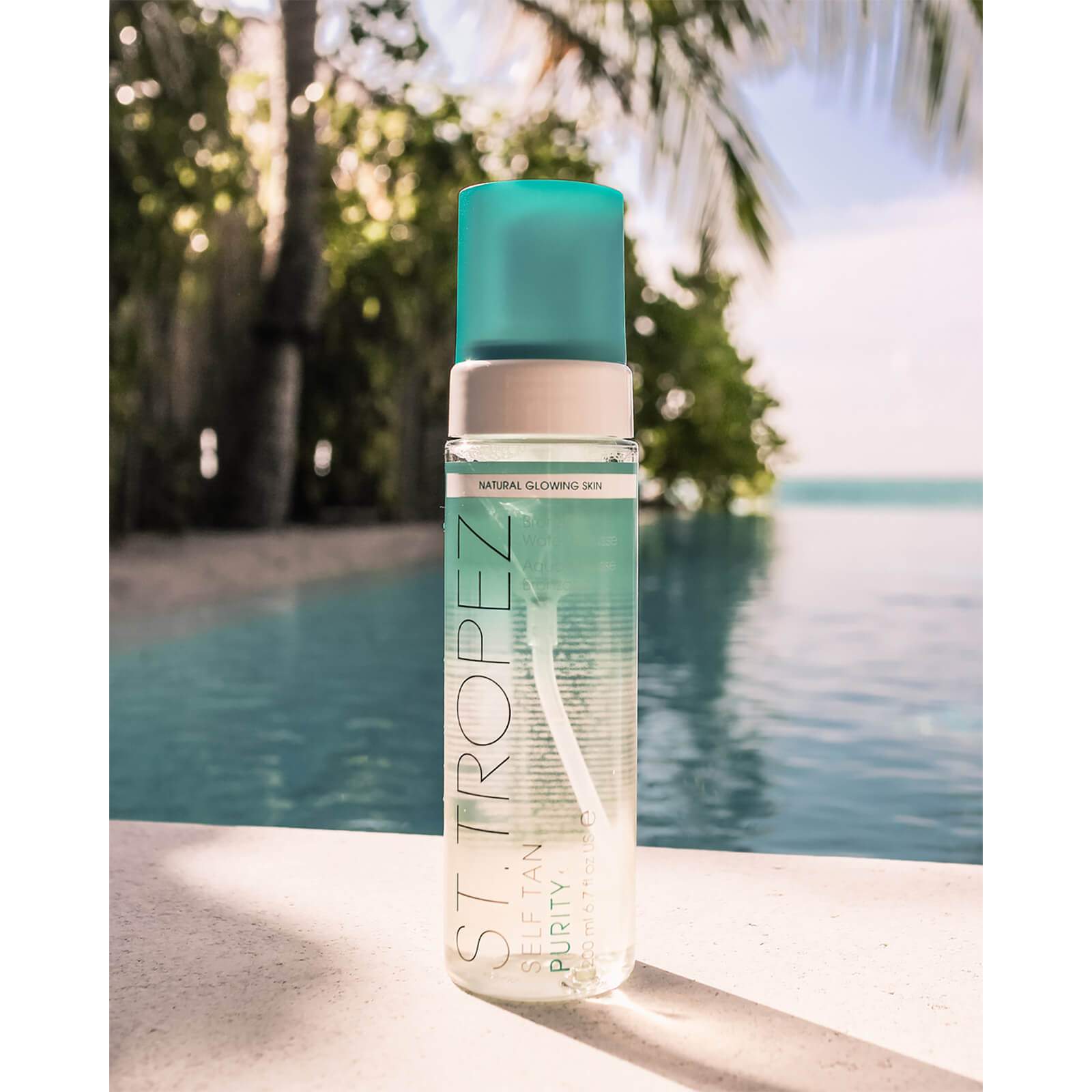 St Tropez self tanning mousse bottle with turquoise cap by poolside and palm trees in background