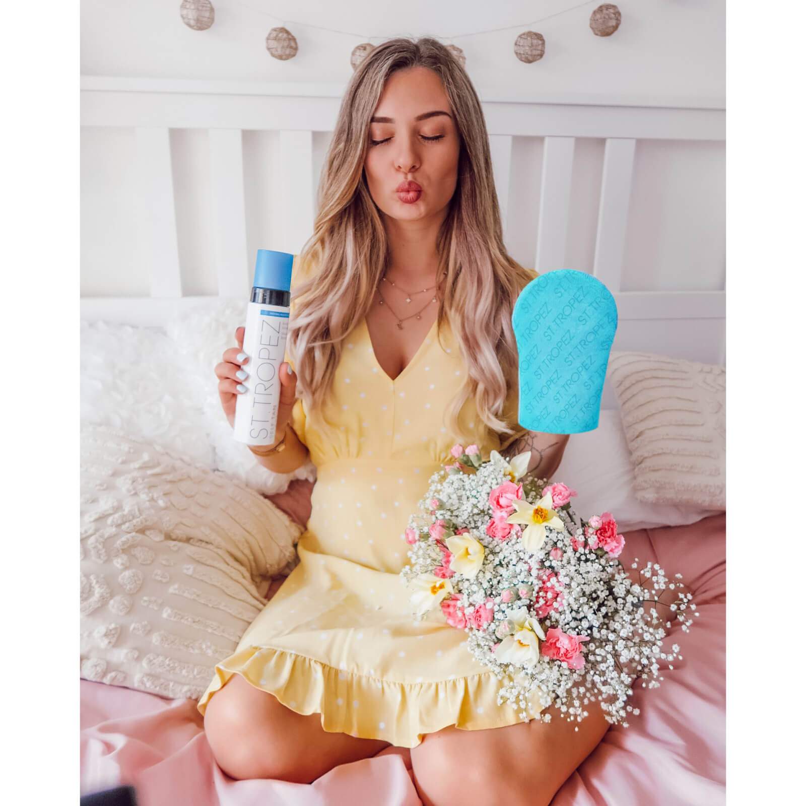 Woman in yellow dress holding St Tropez self tanning mousse and blue applicator mitt while sitting on bed with flowers
