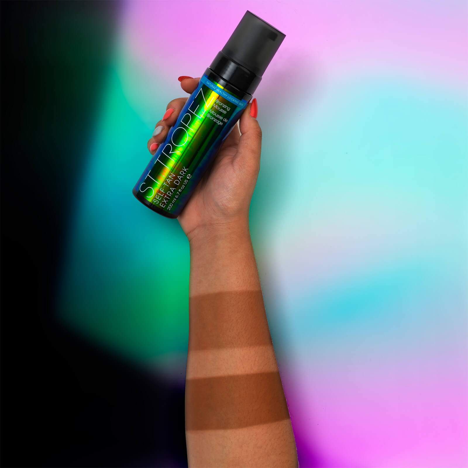 Hand holding St Tropez extra dark bronzing mousse bottle showing gradual tan shades on forearm