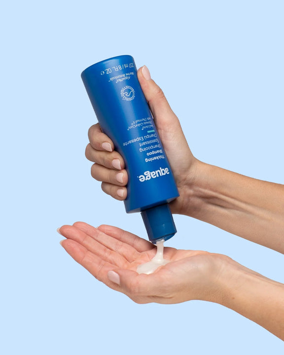 Hand pouring creamy Aquage Thickening Shampoo from blue bottle onto palm over light blue background