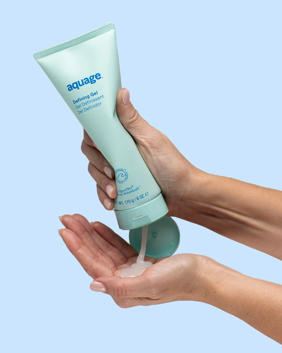 Hands dispensing Aquage Defining Gel from a light blue tube against a light blue background
