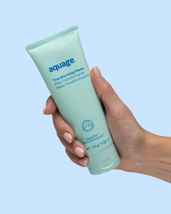 Hand holding a tube of Aquage Transforming Paste hair styling product with aqua blue background