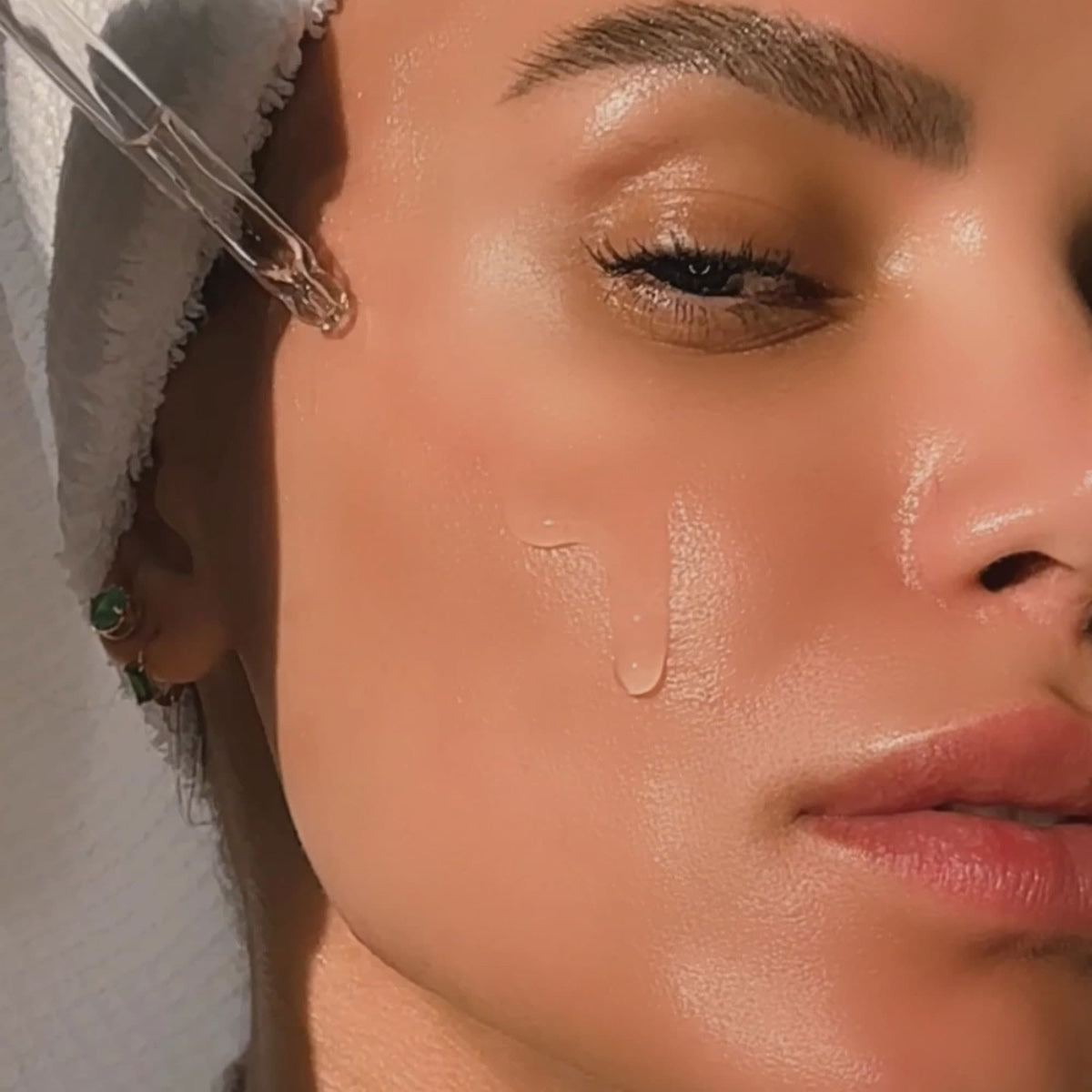 Close up of woman applying Dermelect Rapid Repair Serum with dropper on glowing dewy cheek and towel wrap
