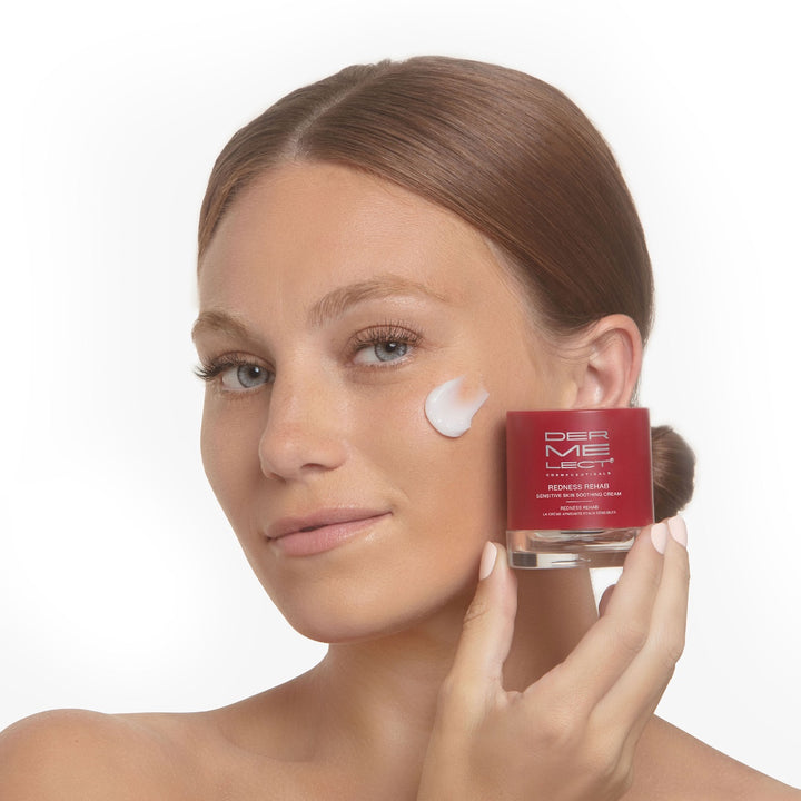 Smiling woman holds Dermelect Redness Rehab cream jar with white moisturizer dab on cheek