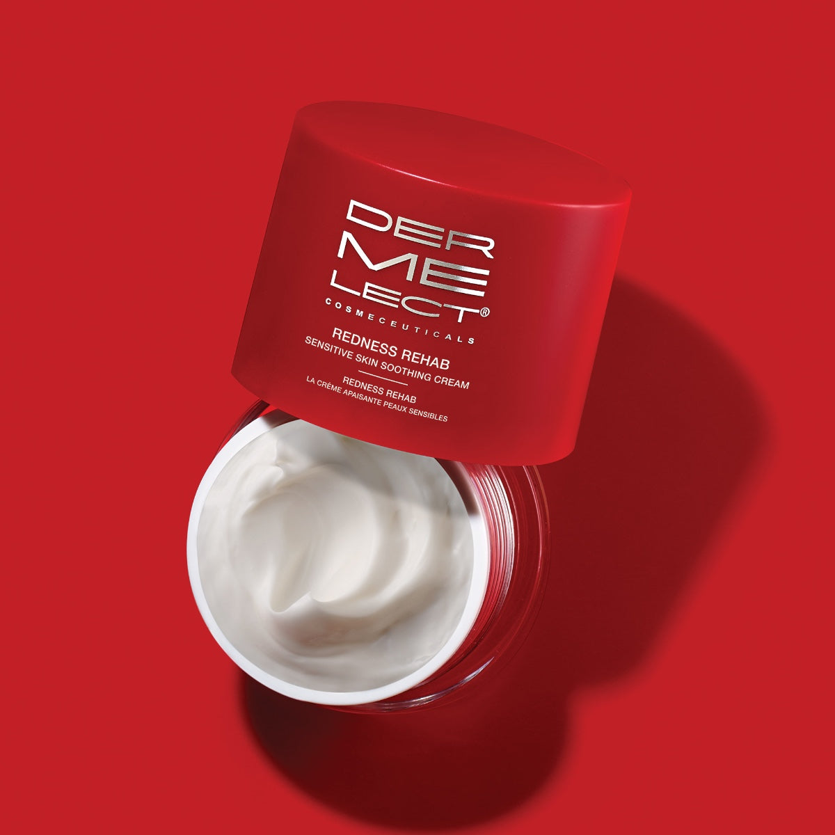 Open red jar of Dermelect Redness Rehab cream showing white soothing moisturizer inside on red background for sensitive skin