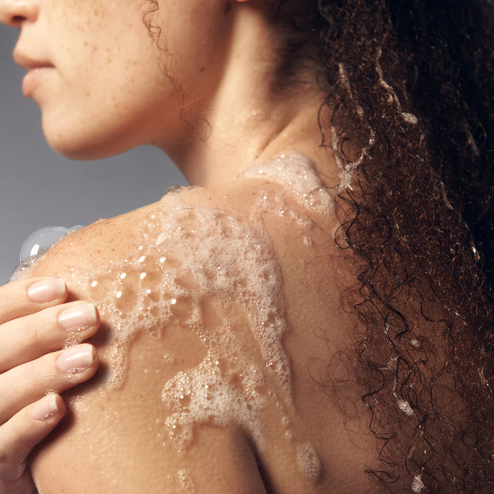 Close-up of a woman applying Philosophy Amazing Grace Jasmine shower gel with foam on shoulder and curly hair