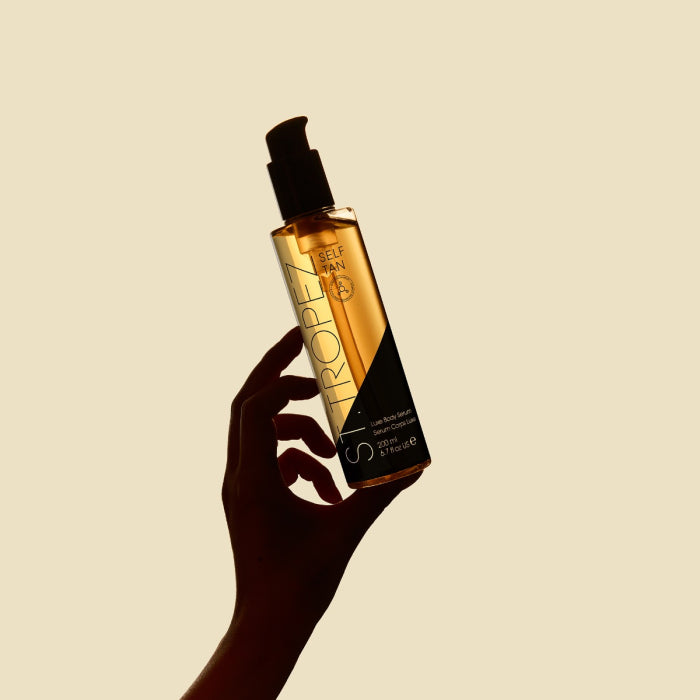 Hand holding a bottle of St Tropez body serum self tan with pump dispenser against beige background