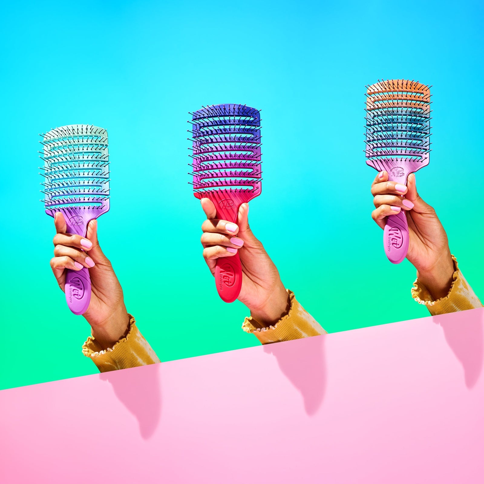 Three colorful Wet Brush Pro Flex Dry Paddle brushes held by hands against blue and pink background