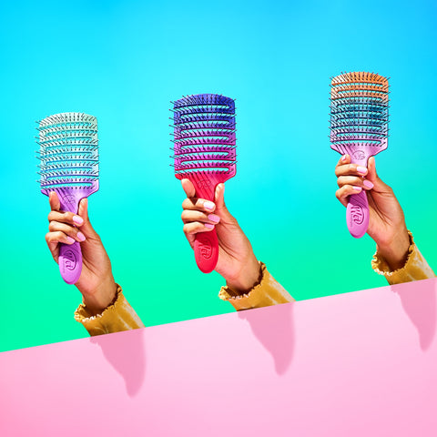 Three colorful Wet Brush Pro Flex Dry Paddle brushes held by hands against blue and pink background