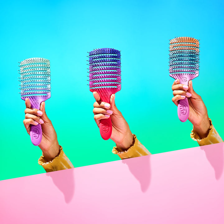 Three colorful Wet Brush Pro Flex Dry Paddle brushes held by hands against blue and pink background