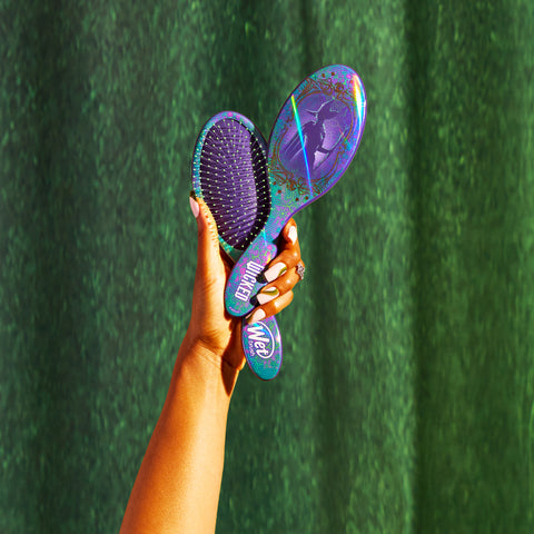 Hand holding Wet Brush Wicked detangling brush with purple and teal design against green background